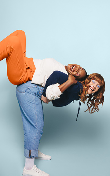 Zwei junge Frauen stehen lachend Rücken an Rücken. Die eine hebt die andere hoch.