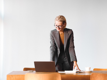 Eine ältere Frau mit Brille stützt sich mit beiden Armen auf einen Tisch und blickt auf ihren Laptop.