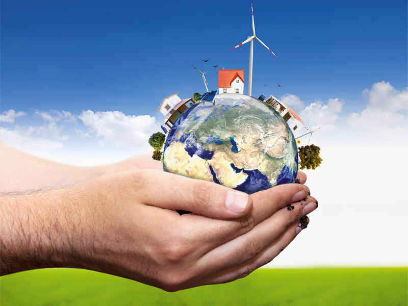 In den mit Erde verschmierten Händen einer Person liegt die Erdkugel auf der drei Wohnhäuser sowie zahlreiche Windräder zu sehen sind.