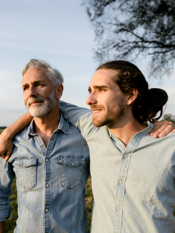 Zwei Männer stehen draußen in der Natur und haben die Arme über die Schulter des anderen gelegt.