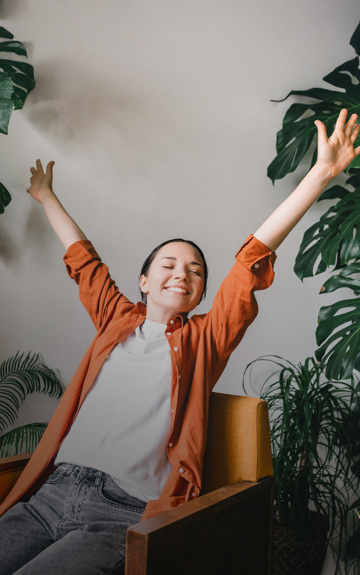 Eine Frau sitzt auf einem Stuhl inmitten von Pflanzen und streckt freudig die Hände in die Höhe.