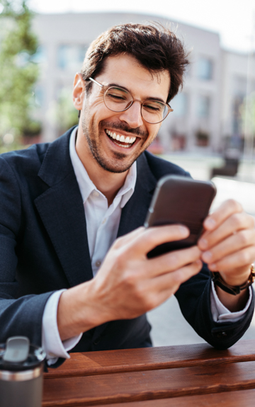 Ein Mann sitzt im Freien auf einer Bank und Blickt mit einem breiten Lächeln auf sein Mobiltelefon.